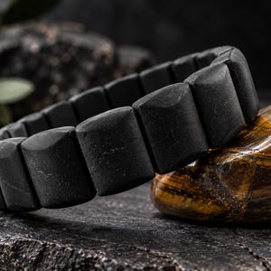 Könnte beinhalten: Ein Armband bestehend aus mattschwarzen, rechteckigen, leicht gewölbten Segmenten. Die einzelnen Teile sind miteinander verbunden und bilden ein durchgehendes Band. Die Oberfläche jedes Segments hat eine glatte, dunkle Textur, was dem gesamten Schmuckstück ein einheitliches Aussehen verleiht.