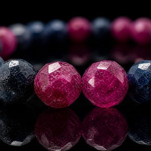 Op de afbeelding: Close-up van een armband met facetgeslepen kralen. De armband wisselt af tussen diepblauwe en levendige magenta kralen. De kralen zijn rond en reflecteren licht, waardoor een sprankelend effect ontstaat. De achtergrond is zwart.