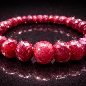Könnte beinhalten: Ein Armband aus facettierten Rubinen. Die tiefroten Edelsteine sind rund und zu einem Kreis aufgereiht. Die Edelsteine haben eine funkelnde, reflektierende Qualität. Das Armband wird auf einer dunklen Oberfläche präsentiert.