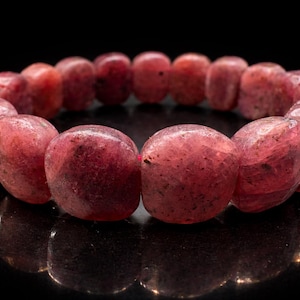 Op de afbeelding: Een close-up van een armband gemaakt van gepolijste, roodroze edelstenen. De armband is cirkelvormig, waarbij elke steen ongeveer 2 cm in diameter is. De achtergrond is zwart, wat de kleur en textuur van de armband benadrukt.