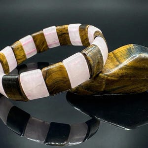 May include: A gemstone bracelet featuring alternating rectangular beads of tiger's eye, rose quartz, and black stone. The bracelet is displayed with a matching tiger's eye stone, all set against a reflective black surface.