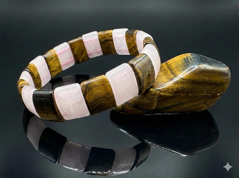 May include: A gemstone bracelet featuring alternating rectangular beads of tiger's eye, rose quartz, and black stone. The bracelet is displayed with a matching tiger's eye stone, all set against a reflective black surface.