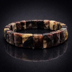 Puede incluir: Una pulsera de cuentas con piedras rectangulares multicolores. Las piedras muestran una mezcla de tonos marrones, negros y beige. La pulsera se exhibe sobre una superficie negra reflectante, resaltando la textura y las variaciones de color de la piedra natural.