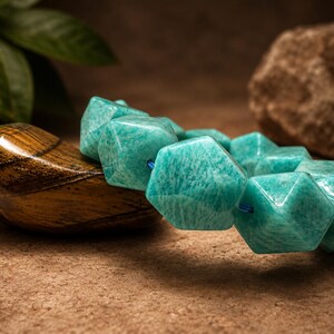 Pu&ograve; includere: Un braccialetto composto da perline esagonali sfaccettate di un vivace colore verde acqua. Le perline sono infilate su un sottile cordino blu, creando un motivo geometrico. La superficie di ogni perlina presenta variazioni naturali di colore.