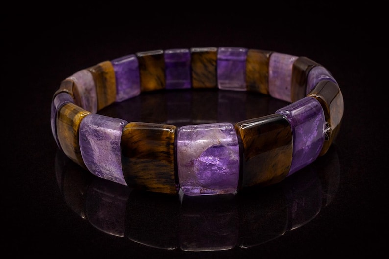 May include: A stretch bracelet featuring alternating rectangular beads of purple and brown tiger's eye stones. The bracelet is displayed on a black surface, reflecting the light.