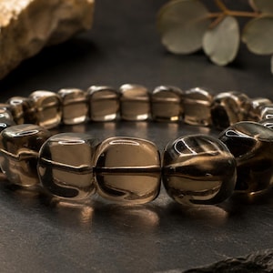Op de afbeelding: Een armband bestaande uit doorschijnende, rookbruine, afgeronde vierkante kralen. De kralen hebben een gepolijste, glanzende afwerking die het licht reflecteert, waardoor er glanspunten op de oppervlakken ontstaan. De kralen vormen een lus.