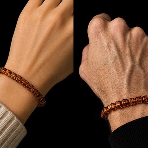 May include: A close-up of two wrists, each adorned with a bracelet. The bracelets are made of amber-colored, square-shaped beads. One wrist is fair-skinned and wearing a white sweater, while the other is darker-skinned and wearing a black long-sleeve shirt.