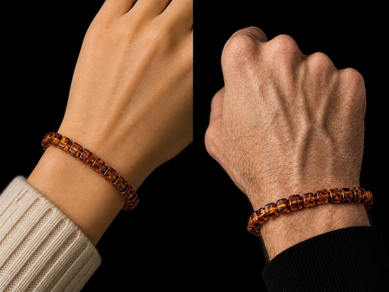 May include: A close-up of two wrists, each adorned with a bracelet. The bracelets are made of amber-colored, square-shaped beads. One wrist is fair-skinned and wearing a white sweater, while the other is darker-skinned and wearing a black long-sleeve shirt.
