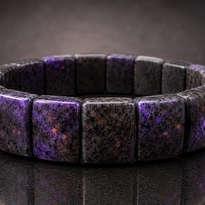 Peut inclure: Un bracelet élastique composé de perles rectangulaires en pierre polie. Les perles présentent un motif moucheté violet foncé et noir avec de subtiles taches orange et dorées sur toute la surface, formant un anneau circulaire continu.