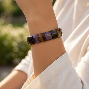 May include: A gemstone bracelet featuring rectangular beads in shades of purple, brown, and black. The bracelet is worn on a wrist, with a white long-sleeved shirt in the background. The bracelet is made of natural stones.