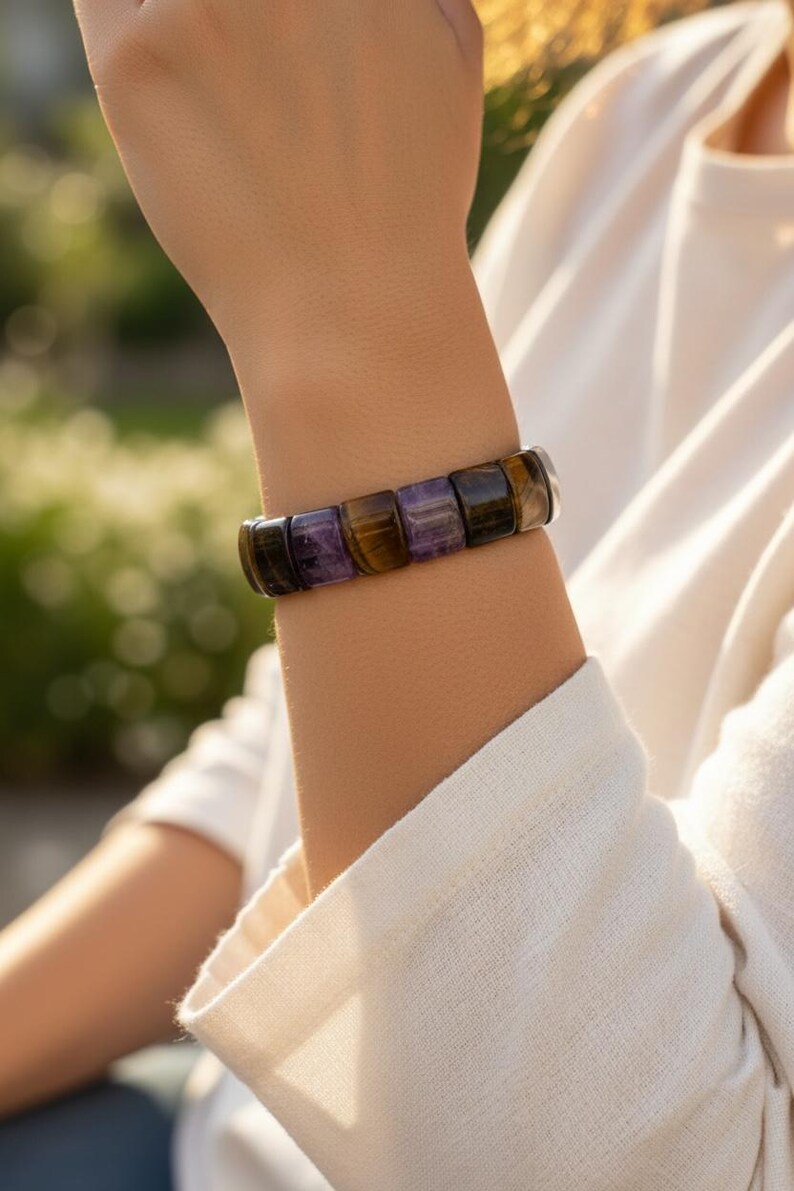 May include: A gemstone bracelet featuring rectangular beads in shades of purple, brown, and black. The bracelet is worn on a wrist, with a white long-sleeved shirt in the background. The bracelet is made of natural stones.