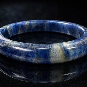 Peut inclure: Un bracelet jonc en pierre précieuse bleue polie. Le bracelet présente un motif marbré de teintes bleu foncé et blanc. Le design lisse et arrondi reflète la lumière, créant un aspect brillant. Le jonc est présenté sur une surface noire réfléchissante.