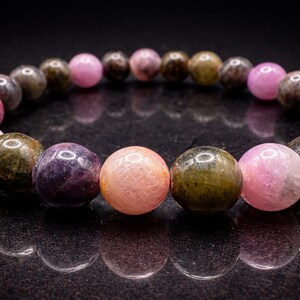 Op de afbeelding: Een armband met ronde edelstenen in verschillende kleuren. De kralen zijn roze, groen, bruin en paars. De armband wordt gepresenteerd op een reflecterend zwart oppervlak.