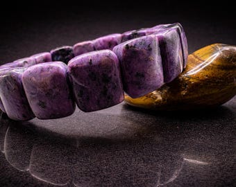 May include: A purple charoite bracelet with rectangular beads rests on a polished, golden-brown tiger's eye stone. The bracelet's color ranges from lavender to deep violet, with dark inclusions. The background is a dark, reflective surface.