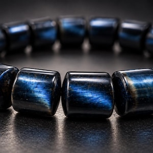 May include: A bracelet composed of cylindrical beads made from dark blue tiger eye stone. Each bead features a polished surface with a chatoyant, shimmering blue lustre that reflects light, arranged in a continuous circular loop pattern.