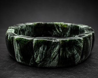 May include: A circular bracelet composed of multiple polished, dark green stone segments. Each segment features a distinct, fibrous, and chatoyant pattern with deep forest green and black tones, creating a continuous, textured, and reflective surface.