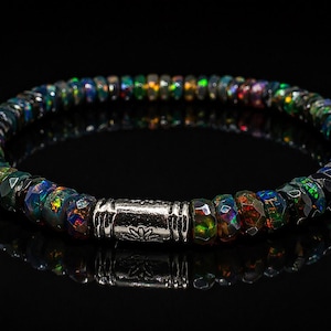 Op de afbeelding: Een kralenarmband met gefacetteerde zwarte opaalstenen met iriserende kleuren. De armband is geregen op een elastisch koord en heeft een zilverkleurige cilindrische sluiting met een bloemmotief. De armband wordt gepresenteerd op een reflecterend zwart oppervlak.