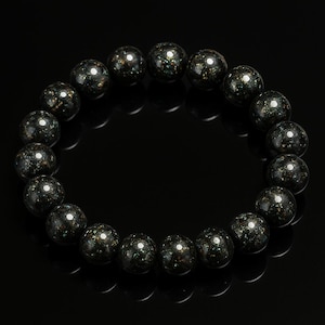 Op de afbeelding: Een armband met donkere, gespikkelde kralen. De kralen zijn zwart met groene en gouden vlekken. De armband is geregen op elastiek, waardoor een cirkel ontstaat. De kralen zijn glad en rond. Elke kraal is ongeveer 1 cm in diameter.