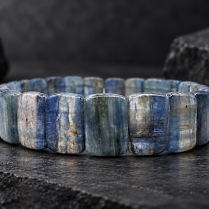 May include: A bracelet composed of rectangular, polished kyanite beads. The beads feature a natural color gradient ranging from deep navy blue to pale greyish-blue, with visible internal striations and variations in the stone texture throughout.