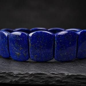 May include: A bracelet composed of polished, rectangular lapis lazuli beads. Each bead features a deep, saturated blue color with natural golden pyrite inclusions scattered across the smooth, glossy surfaces of the stone segments.