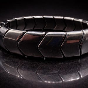 Może przedstawiać: Ciemna, metaliczna bransoletka z wzorem w kształcie szewronu. Bransoletka składa się z połączonych, strzałkowatych ogniw, które tworzą elastyczną opaskę. Powierzchnia jest odblaskowa, a bransoletka leży na ciemnej, teksturowanej powierzchni.