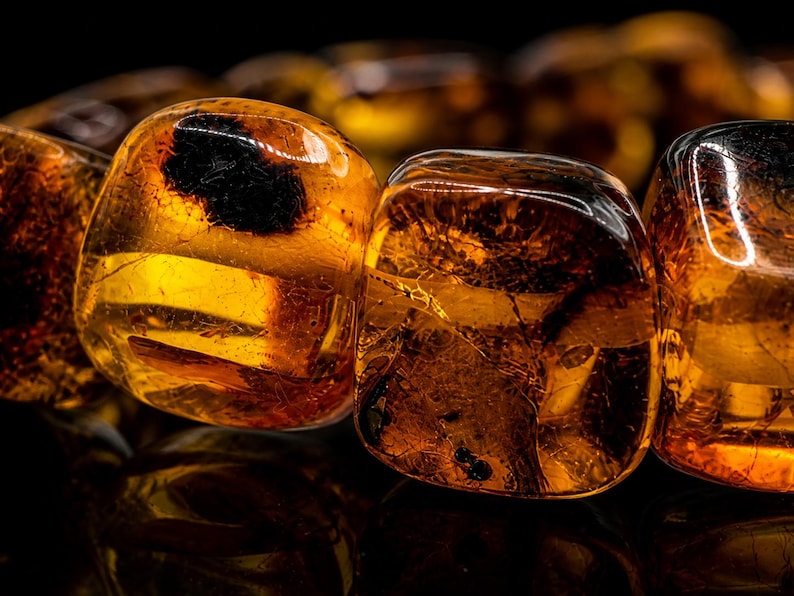 May include: Close-up of polished amber beads, showcasing a warm, translucent golden-brown hue. The beads have a smooth, rounded shape with internal fractures and dark inclusions, creating a unique, natural appearance. The beads are likely part of a necklace or bracelet.