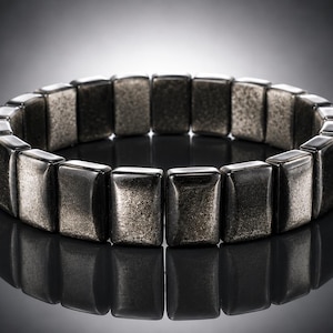 Könnte beinhalten: Ein dunkelgraues Armband mit rechteckigen Gliedern. Die Glieder haben ein strukturiertes, metallisches Aussehen und sind zu einer kreisförmigen Form verbunden. Das Armband wird auf einer reflektierenden Oberfläche präsentiert, wodurch ein Spiegeleffekt entsteht.