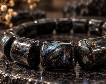May include: A bracelet composed of polished, cylindrical beads made of dark, iridescent stone. The beads feature deep black and brown tones with striking flashes of metallic blue and gold chatoyancy visible across their smooth, reflective surfaces.