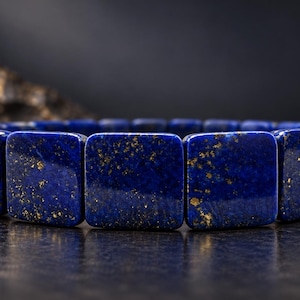 Könnte beinhalten: Ein Armband aus glatten, quadratischen Lapis-Lazuli-Perlen. Jede tiefblaue Perle weist natürliche goldene Pyrit-Einschlüsse auf, die über die Oberfläche verteilt sind und ein einheitliches Muster über die gesamte Länge des Schmuckstücks bilden.