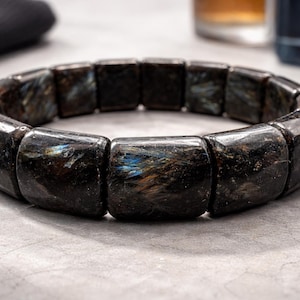 Op de afbeelding: Een donkere armband met rechthoekige kralen. De kralen zijn donker, gespikkeld met hints van blauw en goud. De armband wordt gepresenteerd op een lichtgrijze ondergrond.