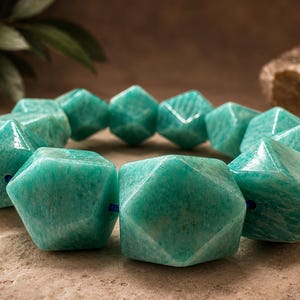 Pu&ograve; includere: Un braccialetto composto da grandi perline geometriche sfaccettate realizzate in pietra amazzonite lucida color verde acqua. Le perline sono infilate su un sottile cordino blu, creando una forma circolare con una finitura liscia e lucida e venature minerali.