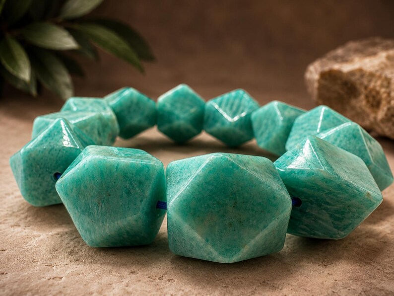 Pu&ograve; includere: Un braccialetto composto da grandi perline geometriche sfaccettate realizzate in pietra amazzonite lucida color verde acqua. Le perline sono infilate su un sottile cordino blu, creando una forma circolare con una finitura liscia e lucida e venature minerali.