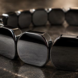 Op de afbeelding: Een close-up van een donkere, glanzende armband. De armband is gemaakt van grote, rechthoekige, gepolijste, zwarte stenen. De stenen zijn verbonden om een cirkelvorm te vormen. De armband ligt op een donkere, getextureerde ondergrond.