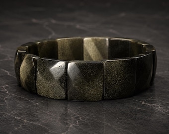 May include: A dark, gold-flecked bracelet composed of rectangular, faceted beads. The bracelet has a rich, earthy tone and a polished finish, reflecting light. The jewelry piece is set against a dark, textured background.