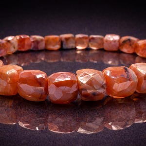Op de afbeelding: Een armband van gefacetteerde oranje edelstenen. De kralen hebben een warme, roodachtig-oranje kleur met een gepolijst, reflecterend oppervlak. De armband wordt getoond op een donkere ondergrond, met zijn reflectie zichtbaar.