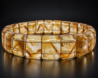 Può includere: Un bracciale composto da perline rettangolari in citrino traslucido color marrone dorato. Ogni perla presenta una superficie lucida con inclusioni naturali interne visibili e striature minerali marrone scuro, disposte in un cerchio continuo.