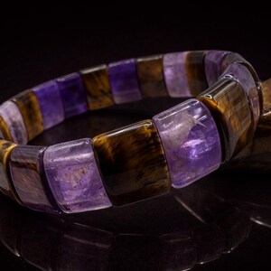May include: A gemstone bracelet featuring alternating rectangular beads of purple amethyst and brown tiger's eye. The bracelet is displayed on a reflective black surface, highlighting the polished stones and their natural patterns. A piece of wood is in the background.