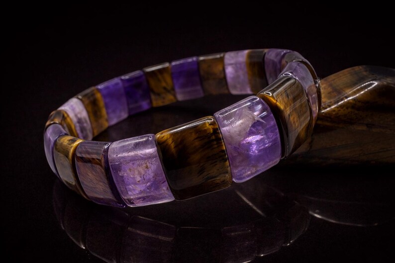 May include: A gemstone bracelet featuring alternating rectangular beads of purple amethyst and brown tiger's eye. The bracelet is displayed on a reflective black surface, highlighting the polished stones and their natural patterns. A piece of wood is in the background.