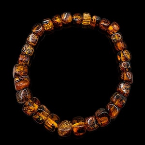 May include: A bracelet made of amber-colored, square-cut beads. The beads have a translucent quality, with variations in color and texture, ranging from light honey to deep brown. The bracelet is set against a black background.