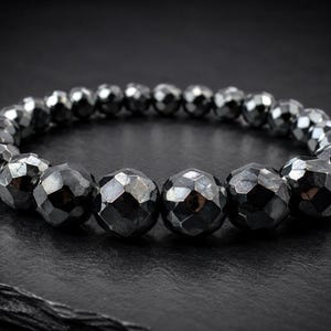 Op de afbeelding: Een elastische armband bestaande uit talloze donkergrijze, metallic, gefacetteerde bolvormige kralen. De kralen hebben een reflecterend, gepolijst oppervlak met meerdere platte geometrische vlakken die het licht vangen, wat zorgt voor een getextureerd uiterlijk.