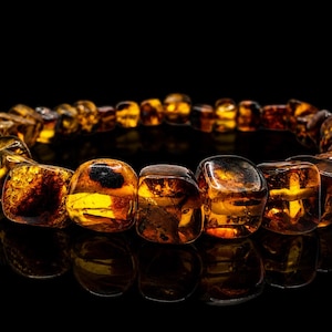 May include: A bracelet made of amber beads. The beads are a mix of golden and brown hues, with a polished, reflective surface. The bracelet is set against a black background, highlighting the amber's natural colors and textures.