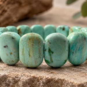 Könnte beinhalten: Ein elastisches Armband aus glatten, oval geformten, türkis farbenen Perlen. Jede Perle weist natürliche Farbvariationen auf, darunter Schattierungen von Hellblau, Grün und braune Matrixeinschlüsse, die in einem Kreis angeordnet sind.
