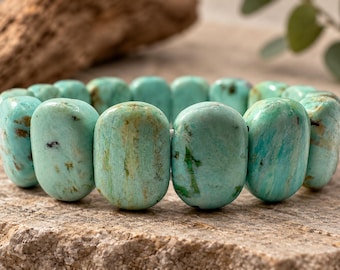 May include: A stretch bracelet composed of smooth, oval-shaped turquoise-colored beads. Each bead features natural variations in color, including shades of light blue, green, and brown matrix inclusions, arranged in a continuous circular pattern.