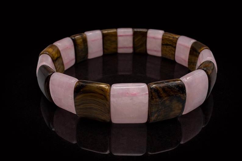 May include: A bracelet featuring alternating rectangular beads of pink and brown tiger eye stone. The bracelet is displayed on a black surface, showcasing its polished finish and natural color variations. The jewelry is a fashion accessory.