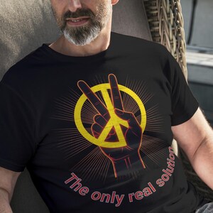 Puede incluir: Camiseta negra con un símbolo de la paz amarillo y el texto "The only real solution" en rojo. El diseño presenta una mano haciendo el signo de la paz. La persona está sentada en una silla de mimbre.