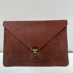 Könnte beinhalten: Eine braune Laptop-Hülle aus Leder im Umschlagstil mit einem messingfarbenen Verschluss. Die Hülle hat eine strukturierte Oberfläche und einen Klappenverschluss. Das Leder weist natürliche Farb- und Texturvariationen auf.