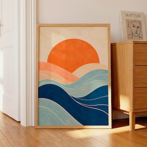 Può includere: Una stampa artistica incorniciata con un paesaggio astratto con un sole arancione che tramonta su onde blu e pesca. L'opera d'arte è in una cornice di legno naturale e si appoggia a una porta bianca e a un comò in legno.