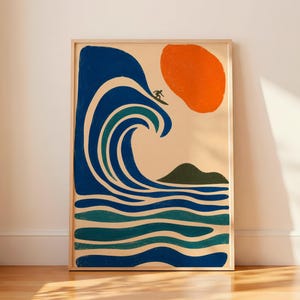 Puede incluir: Una impresión artística enmarcada que presenta una ilustración estilizada de un surfista surfeando una gran ola azul bajo un sol naranja. La obra de arte incluye una pequeña isla verde y está ambientada sobre un fondo crema. El marco es de madera clara.