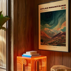May include: A framed art print titled "Atlas Mountains North Africa" with a landscape scene of mountains and a starry sky. The print is displayed above a clear orange acrylic side table with books on top. A wooden stump is next to the table.