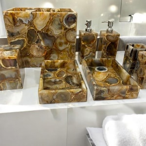 Puede incluir: Un juego de baño que incluye un cesto de basura, dispensadores de jabón, bandejas y porta cepillos de dientes. El juego está hecho de un material de piedra natural con tonos marrones, beige y crema. Las piezas están dispuestas sobre una superficie blanca.